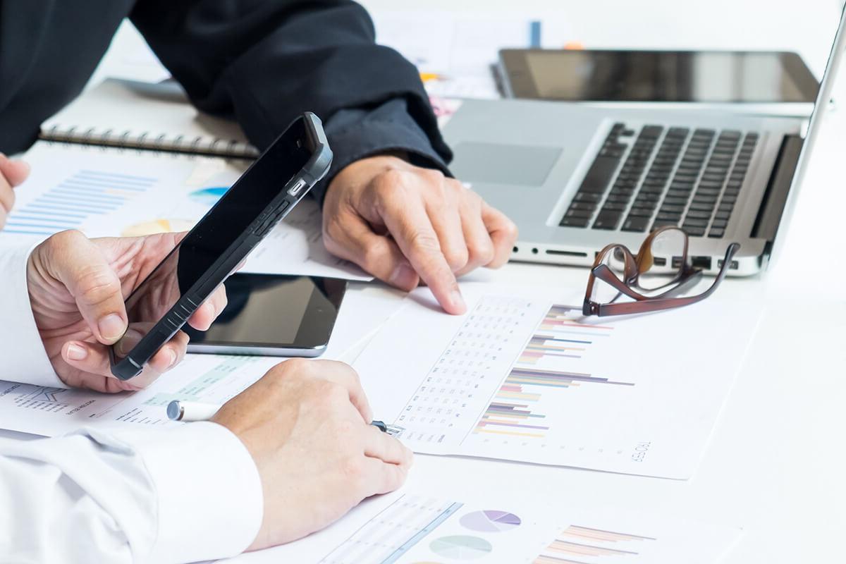 关闭 up of two people's hands holding smartphones and pointing at charts printed out on paper laying on the desk